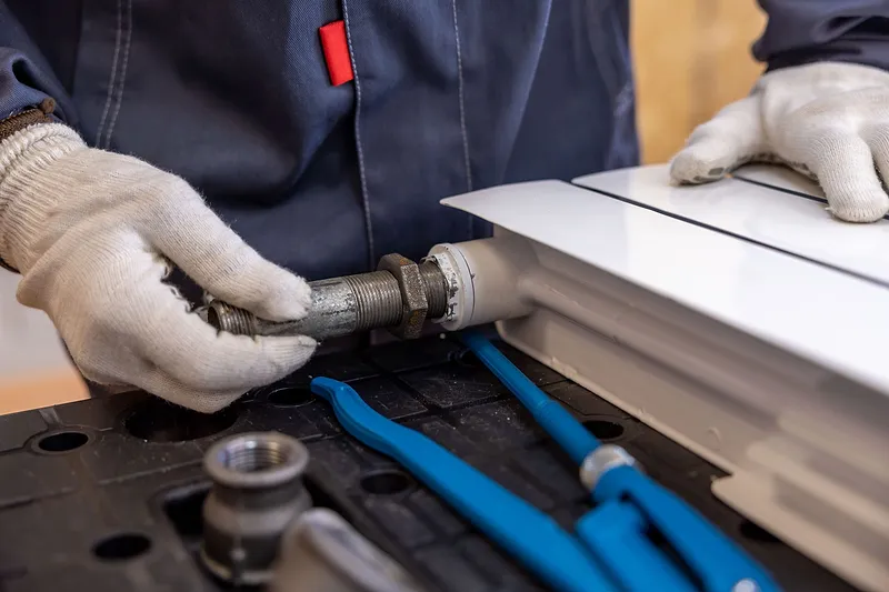 Handwerker mit Arbeitshandschuhen montiert einen Rohranschluss an einem weißen Heizkörper, daneben liegt eine blaue Rohrzange